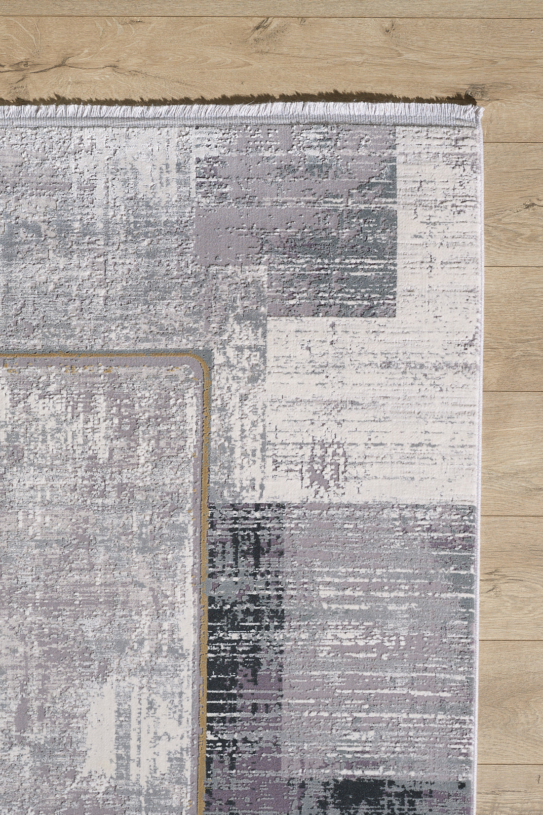 The GridWave Silver Premium Rug - M426A, a modern machine-made design with abstract block patterns in gray, white, and purple plus a short white fringe along the top edge, sits on a light wood floor for a minimalist look.