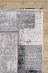 The GridWave Silver Premium Rug - M426A, a modern machine-made design with abstract block patterns in gray, white, and purple plus a short white fringe along the top edge, sits on a light wood floor for a minimalist look.