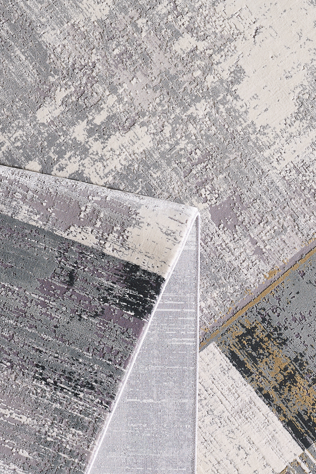 A close-up of the GridWave Silver Premium Rug - M426A, featuring a modern abstract pattern in gray, cream, and touches of gold and lavender. One corner is folded to show the underside. Crafted from acrylic-viscose in neutral tones.