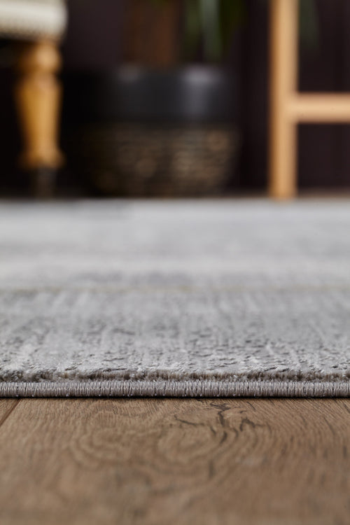 Load image 1 in gallery view. Close-up of the GridWave Silver Premium Rug - M426A, featuring a soft gray acrylic-viscose texture on a wooden floor; blurred furniture and a black planter appear in the background.