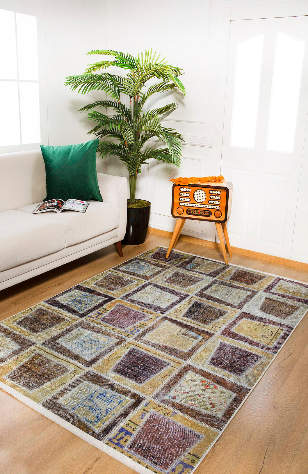 A bright living room features the Gridiron Glow - Washable Rug - JR183 (Custom Size), a white sofa with a green pillow and open magazine, a potted plant, and a vintage-style radio—ideal for high-traffic areas near white double doors.