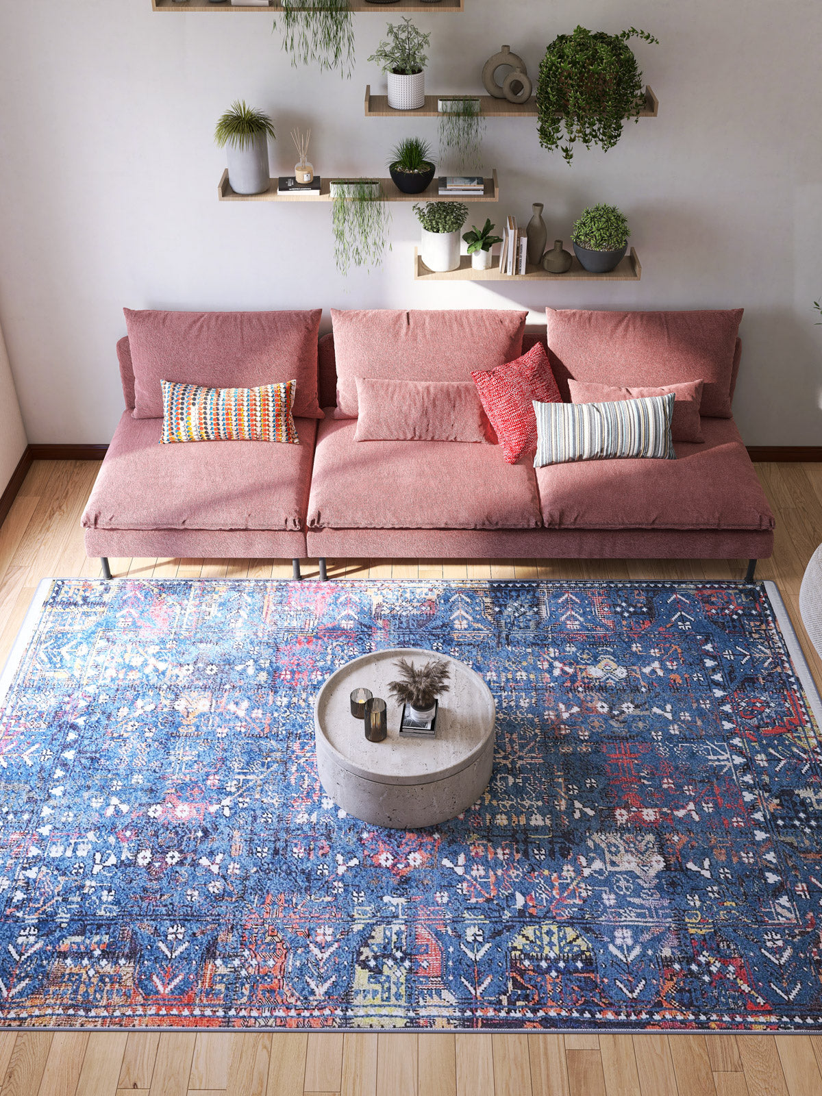 A modern living room with a pink sectional sofa, colorful pillows, and a round concrete coffee table. The space is anchored by the Gypsy Glitz - Washable Rug - JR1650, ideal for high-traffic areas. Shelves of green plants are mounted above the sofa.