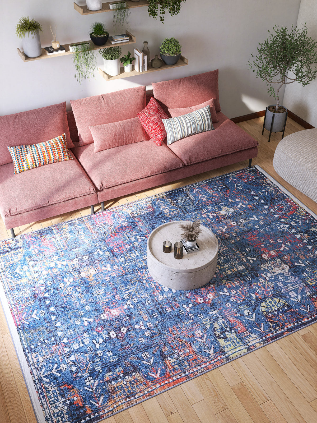 A bright living room features a pink sectional sofa, colorful cushions, the Gypsy Glitz - Washable Rug - JR1650 in blue for high-traffic areas, a round coffee table, wooden floors, potted plants, and wall-mounted shelves with greenery.
