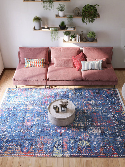 Load image 1 in gallery view. A modern living room with a pink sectional sofa, colorful cushions, a round stone coffee table, the Gypsy Glitz - Washable Rug - JR1650 (Custom Size) ideal for high-traffic areas, plus plants on wall-mounted shelves against a white wall.