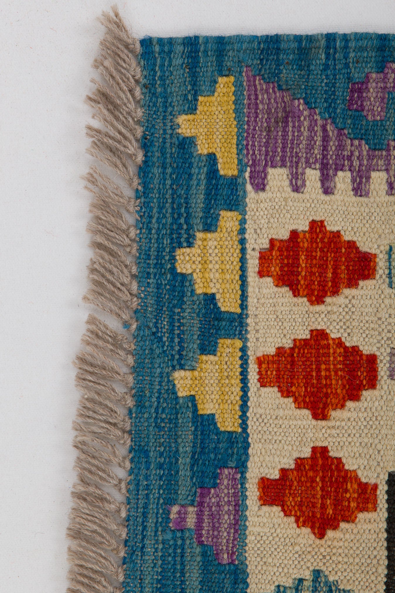 The Handcrafted Story - Handwoven Kilim is a flatweave rug with blue edges, beige fringe, and geometric yellow, purple, orange, and red triangles on a cream and blue background.