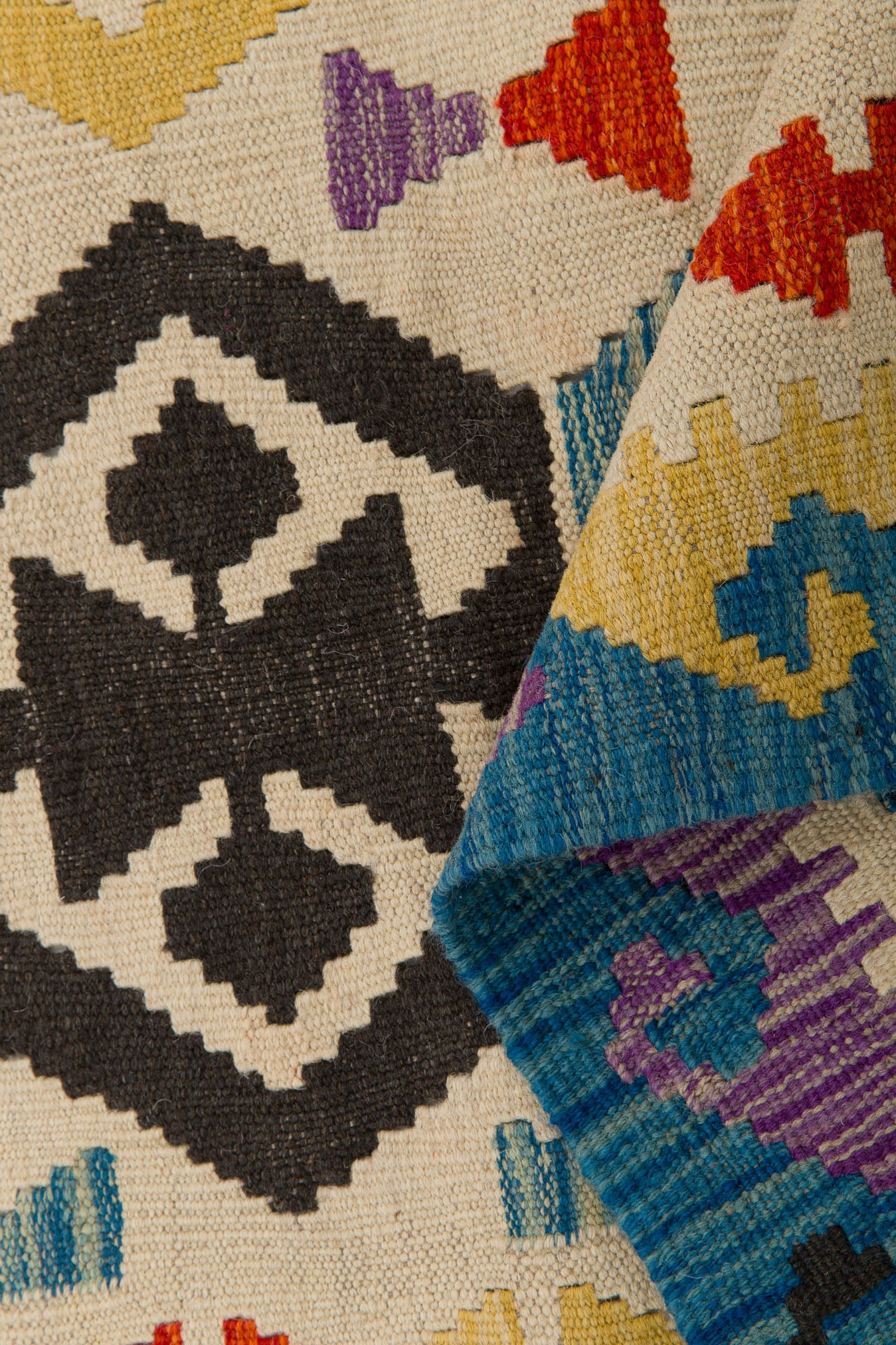 A close-up of the Handcrafted Story - Handwoven Kilim displays black, yellow, blue, red, and purple geometric patterns on a cream wool background; a folded corner reveals colorful zigzag designs on the reverse.