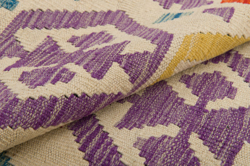 Load image 1 in gallery view. Close-up of the Handcrafted Story - Handwoven Kilim, showcasing its geometric pattern in purple, white, yellow, with touches of red and blue. The folded wool fabric highlights the textile's rich texture and intricate craftsmanship.