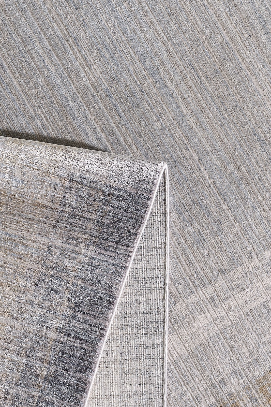 A close-up of the Harmony in Contours Modern Bordered Rug - EW5743 highlights its neutral tone and subtle diagonal stripes, with one corner folded to reveal a lighter-hued underside featuring a similar pattern.