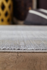A close-up, low-angle view of the Harmony in Contours Modern Bordered Rug - EW5743 in light gray on a wooden floor; the blurred background shows a yellow decor piece and a striped cushion.