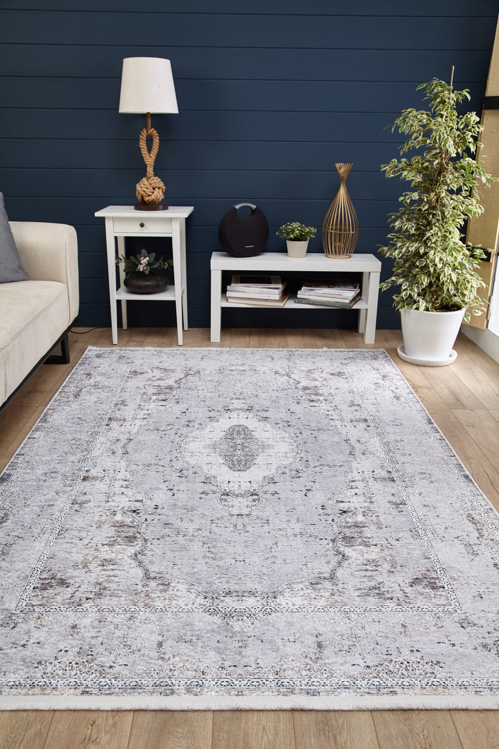 A living room features the Heritage Jewel - Washable Rug - JR1778 (Custom Size) in gray-anthracite on a wood floor, paired with a white couch, two white side tables with books and decor, a potted plant, and a blue paneled wall.