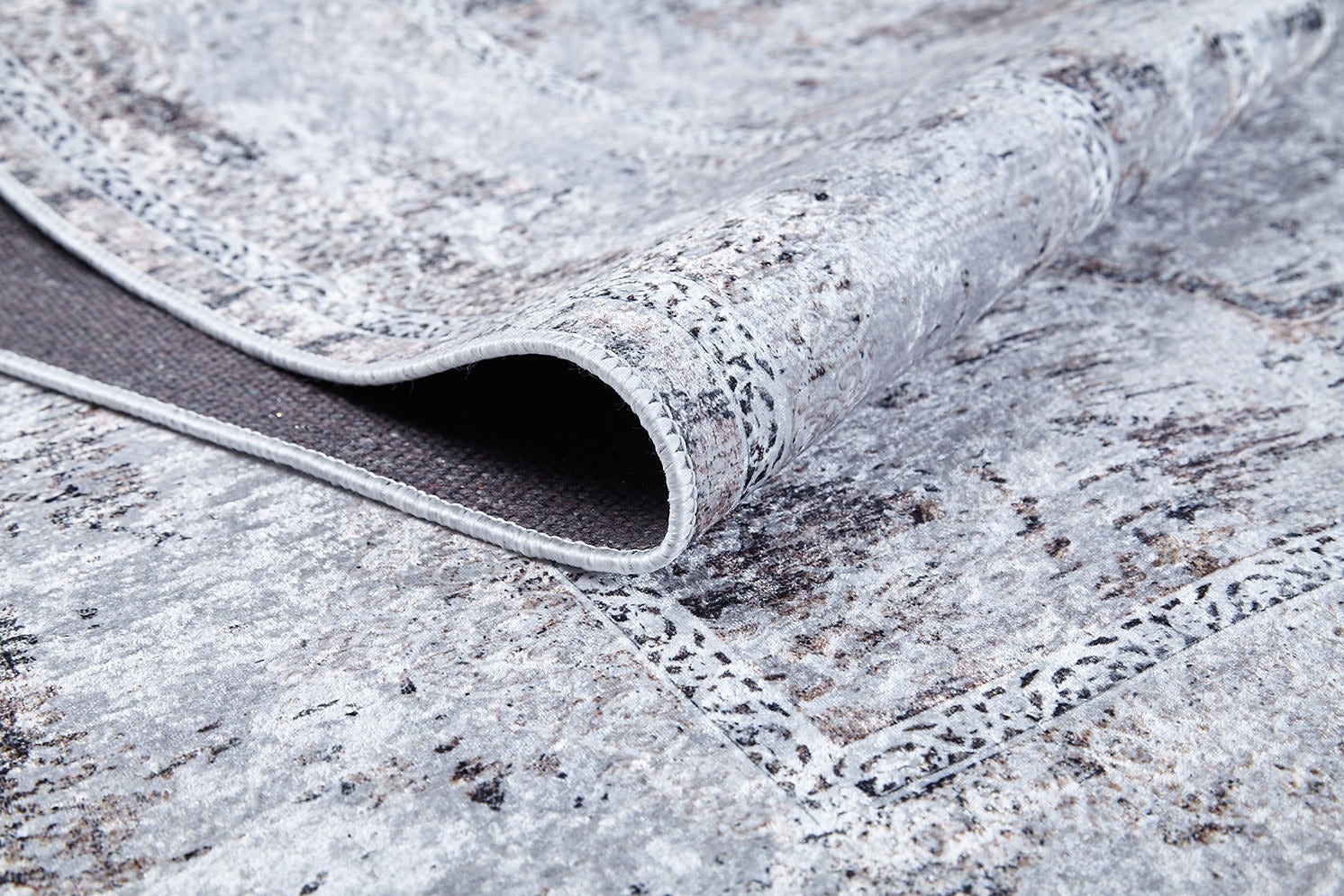 A close-up of the Heritage Jewel - Washable Rug - JR1778 (Custom Size) in gray-anthracite shows its abstract, textured pattern. A folded corner reveals the darker, non-slip backing—ideal for high-traffic spaces.