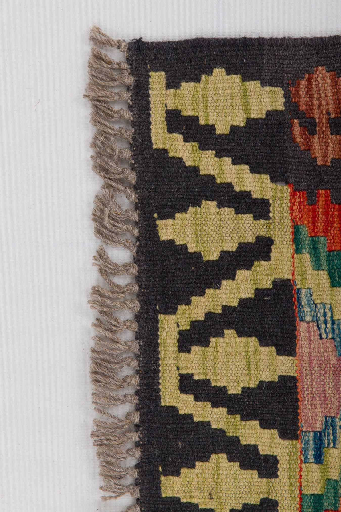 Close-up of the Heritage Weave - Handwoven Kilim rug corner with beige fringe, displaying geometric patterns in green, yellow, black, red, and blue on a white background.