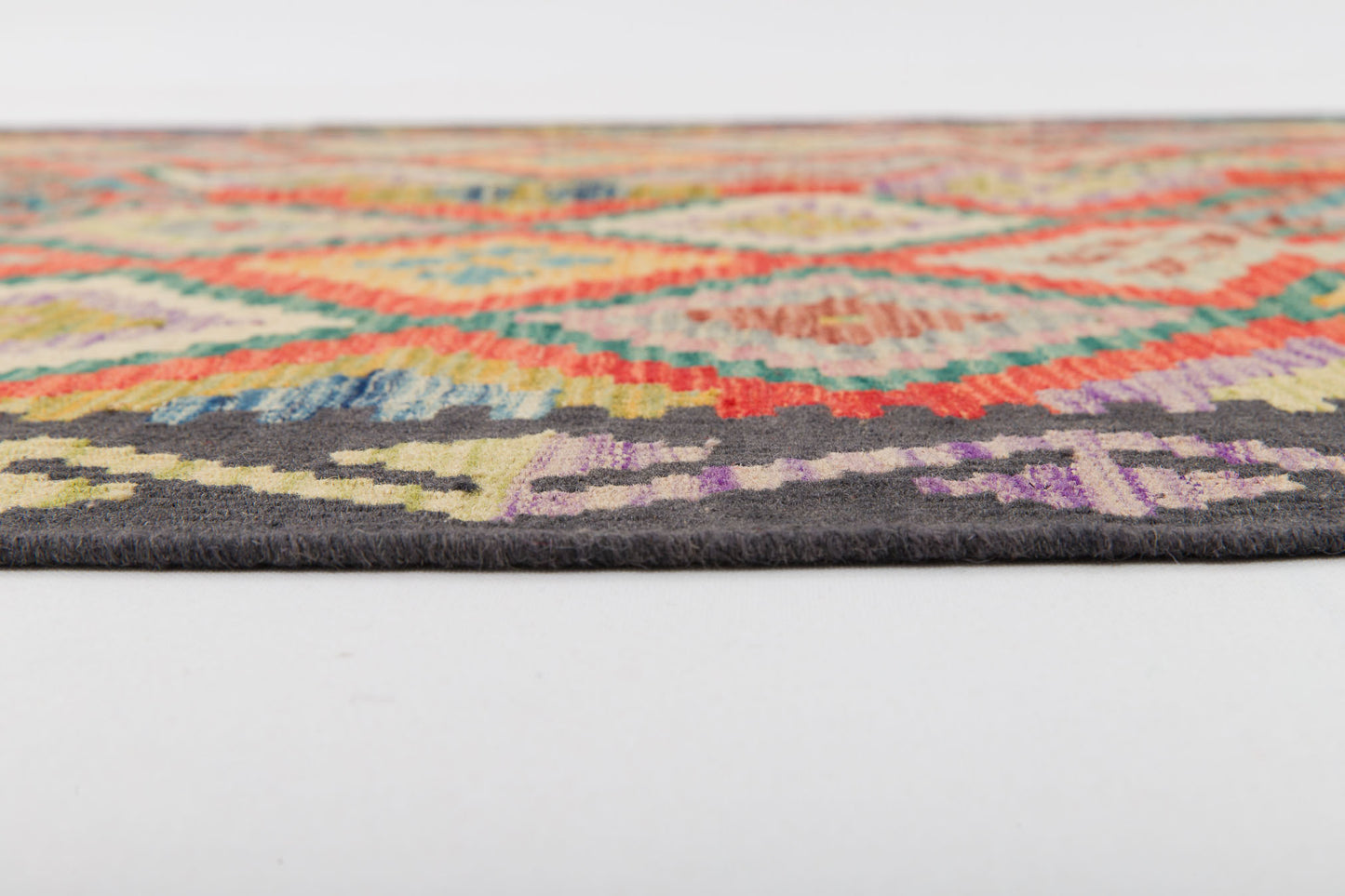 Close-up of the Heritage Weave - Handwoven Kilim, a flat-woven wool rug with a colorful geometric pattern and dark border. This intricate design features vibrant red, yellow, green, blue, and purple shades on a white surface.