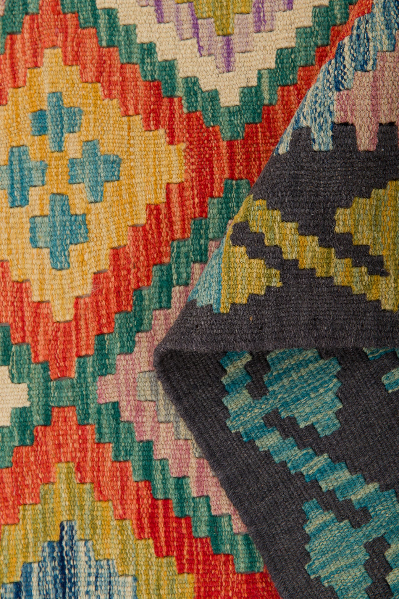 The Heritage Weave - Handwoven Kilim is a colorful flatweave rug featuring geometric patterns in red, yellow, teal, purple, and blue. A folded corner reveals its textured reverse side with classic kilim designs in deeper hues.