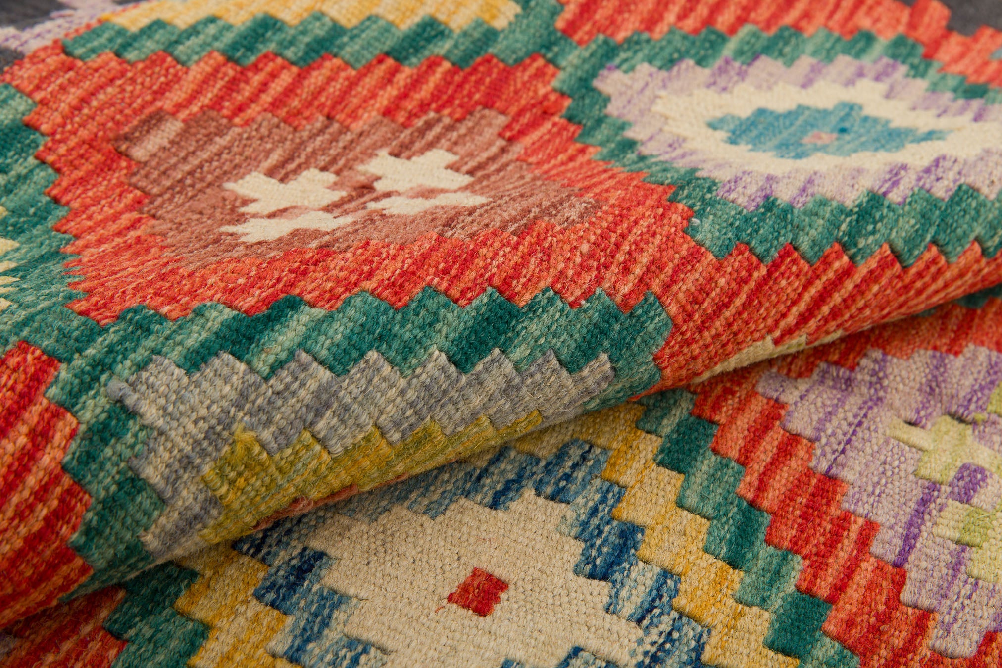 A close-up of the Heritage Weave - Handwoven Kilim shows colorful geometric patterns in red, green, yellow, blue, and white with zigzag and diamond shapes. The folded wool rug highlights its vibrant handwoven texture.