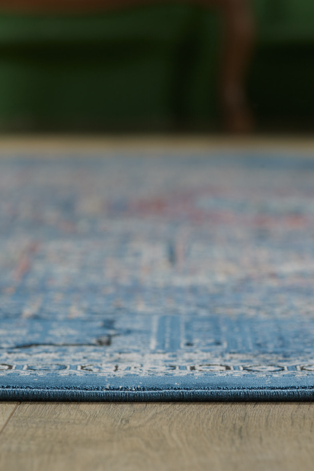 Close-up of the edge of the Hidden Treasures Turkish Rug - Azure - 2223B on a wooden floor, with a blurred green background above.