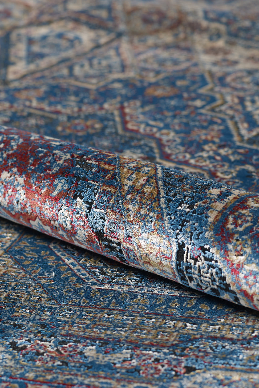 A close-up of the Hidden Treasures Turkish Rug - Azure - 2223B, rolled up to showcase its intricate red, gold, beige, and white patterns. The detailed texture highlights its luxurious and ornate machine-made design.
