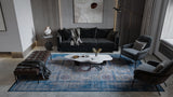 A modern living room features a dark sofa, armchair, bench, and the Hidden Treasures Turkish Rug - Azure - 2223B. A marble coffee table, abstract wall art, floor lamp, and decorative objects complete the cozy, stylish space.