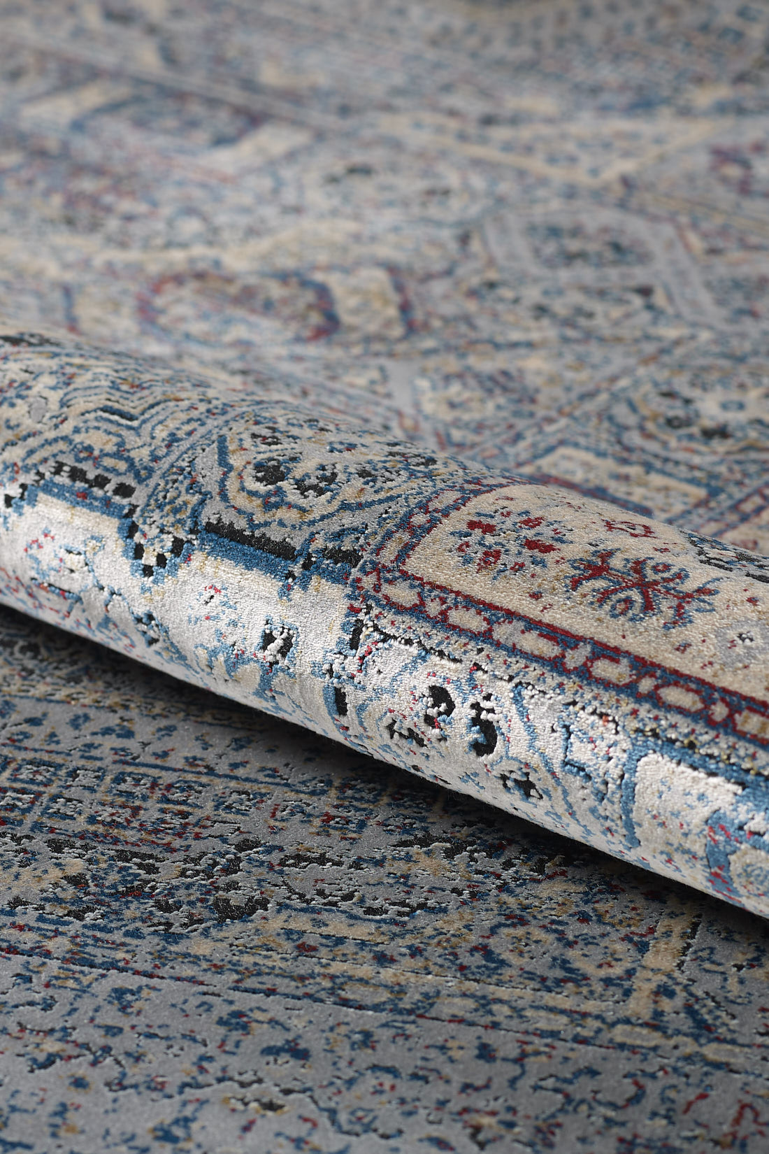 A close-up of the Hidden Treasures Turkish Rug - Smoky - 2223C, showcasing intricate blue, gray, white, and red designs with ornate details and a slightly distressed texture.