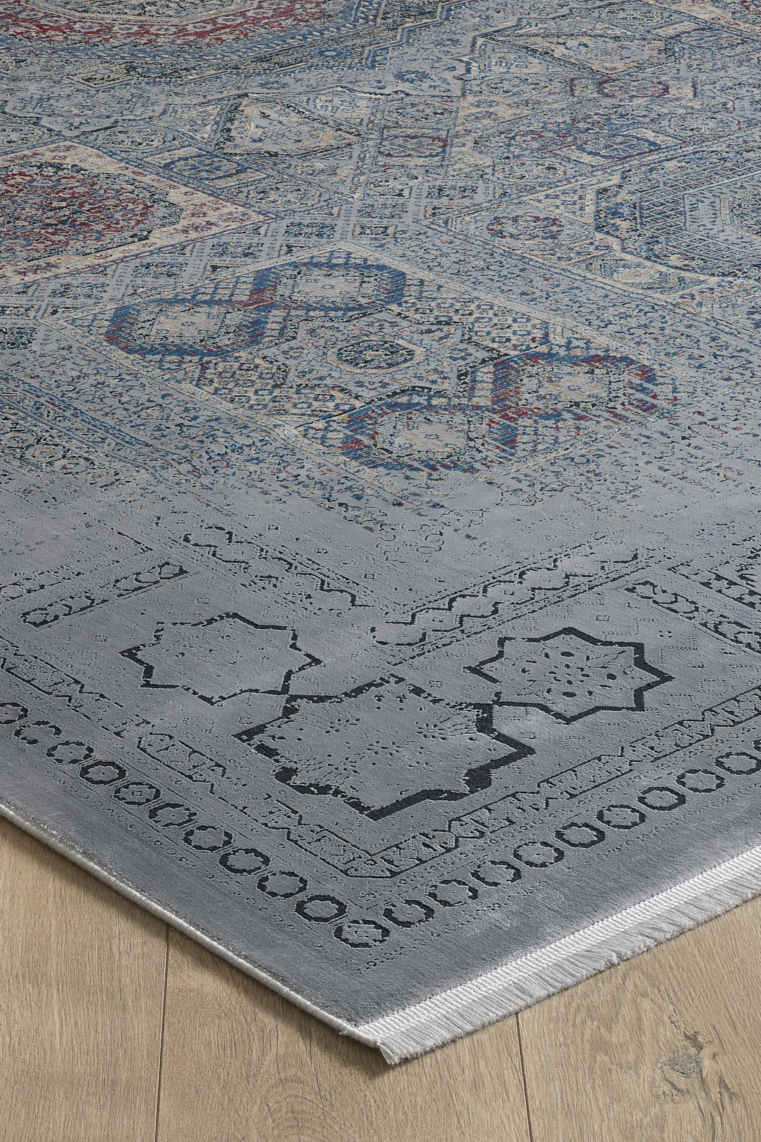 A close-up of the Hidden Treasures Turkish Rug - Smoky - 2223C shows its intricate geometric and floral patterns in dark and light tones, machine-made with fringed edges and detailed textures, placed on a light wood floor.