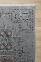 A partial view of the Hidden Treasures Turkish Rug - Smoky - 2223C, featuring geometric and decorative motifs in muted gray, blue, and beige on a light wood floor. Visible fringe accents the top edge of this patterned modal silk rug.