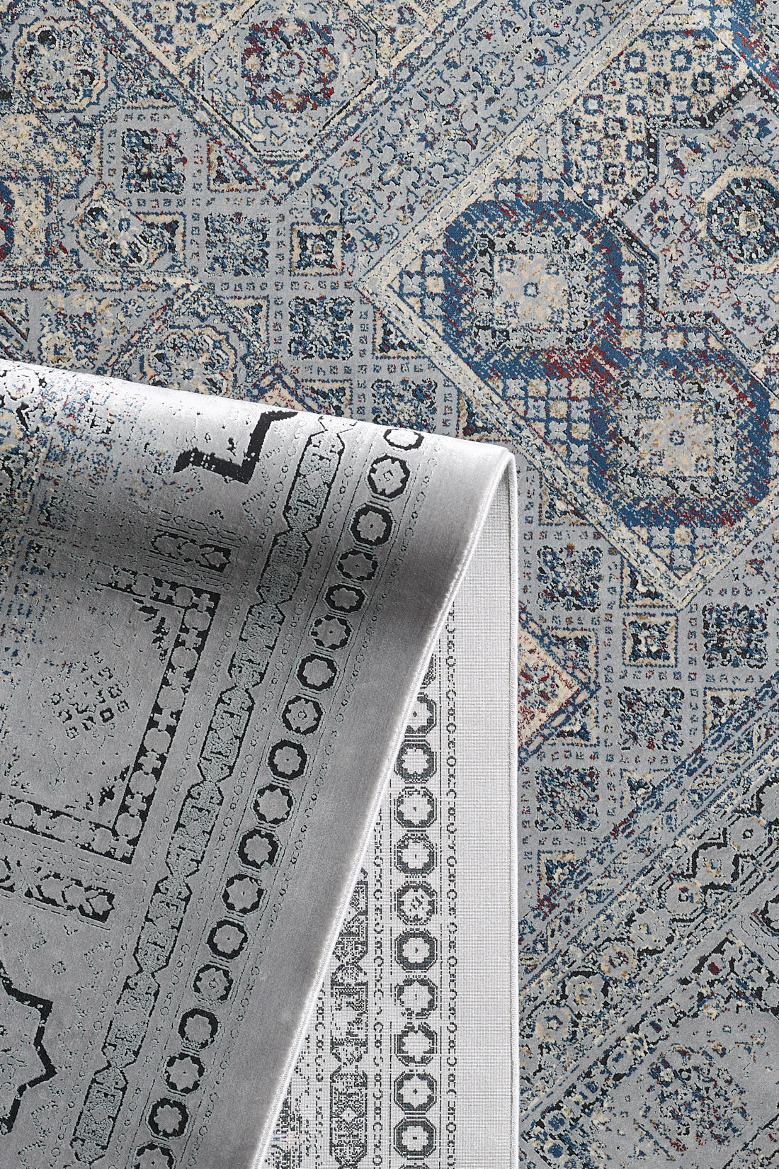 A close-up of the Hidden Treasures Turkish Rug - Smoky - 2223C, featuring blue, grey, and beige geometric and floral designs. One corner is folded over, showing the underside with a faded version of the pattern.
