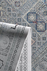 A close-up of the Hidden Treasures Turkish Rug - Smoky - 2223C, featuring blue, grey, and beige geometric and floral designs. One corner is folded over, showing the underside with a faded version of the pattern.