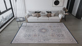 A modern living room features a light sofa with cushions, round side tables, white curtains on a large window, and abstract art above. The Hidden Treasures Turkish Rug - Smoky - 2223C adds patterned style underfoot.