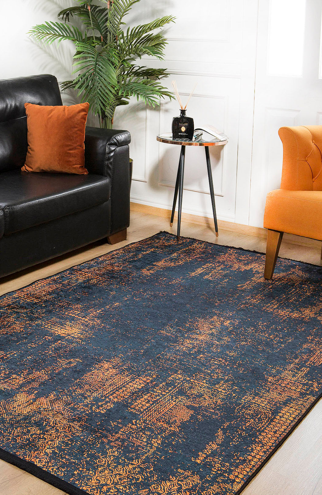 A cozy living room with a black sofa, orange pillow, orange armchair, the Hushed Tones - Washable Rug - JR1428 (Custom Size) in dark blue and orange for high-traffic areas, a small round side table with decor, and a potted plant in the corner.