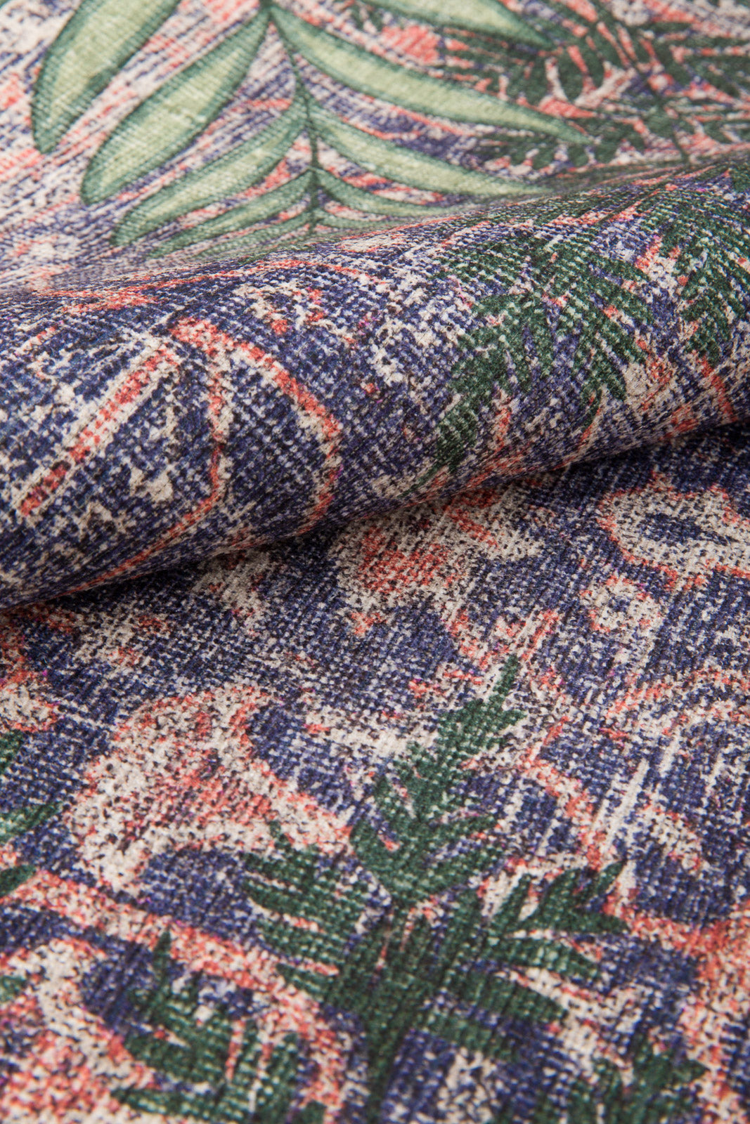 A close-up of the Stormstripe Echo - Washable Rug (JR0046), showing its thick, woven texture and multicolored pattern with green leaves and abstract shapes in blue, red, and cream. The fabric is slightly folded to highlight the weave detail.