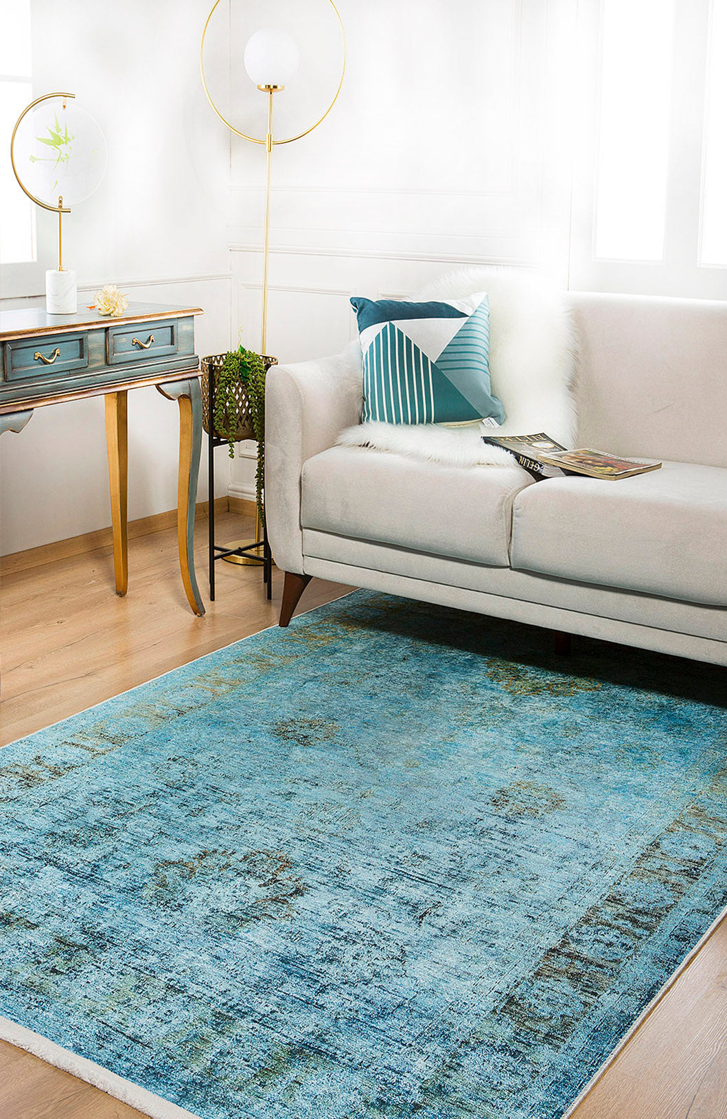 A modern living room features a light beige sofa, blue patterned pillow, glass-top desk with drawers, gold floor lamp, and an allergy-sensitive Imperial Jewel - Washable Rug - JR162 (Custom Size) on wood floors. A magazine rests on the sofa; a plant sits on the desk.
