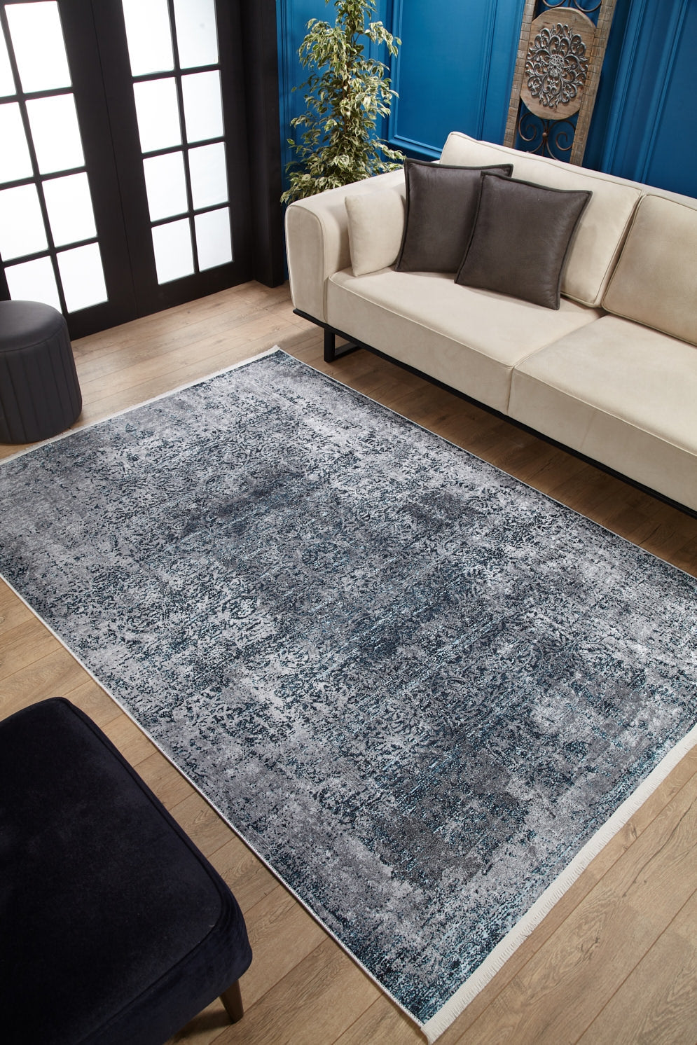 A modern living room features a beige sofa, dark accent pillows, the Imperial Lace - Washable Rug - JR1607 (Custom Size) in blue for high-traffic areas on a light wood floor, a dark ottoman, a plant, and blue paneled walls with décor.