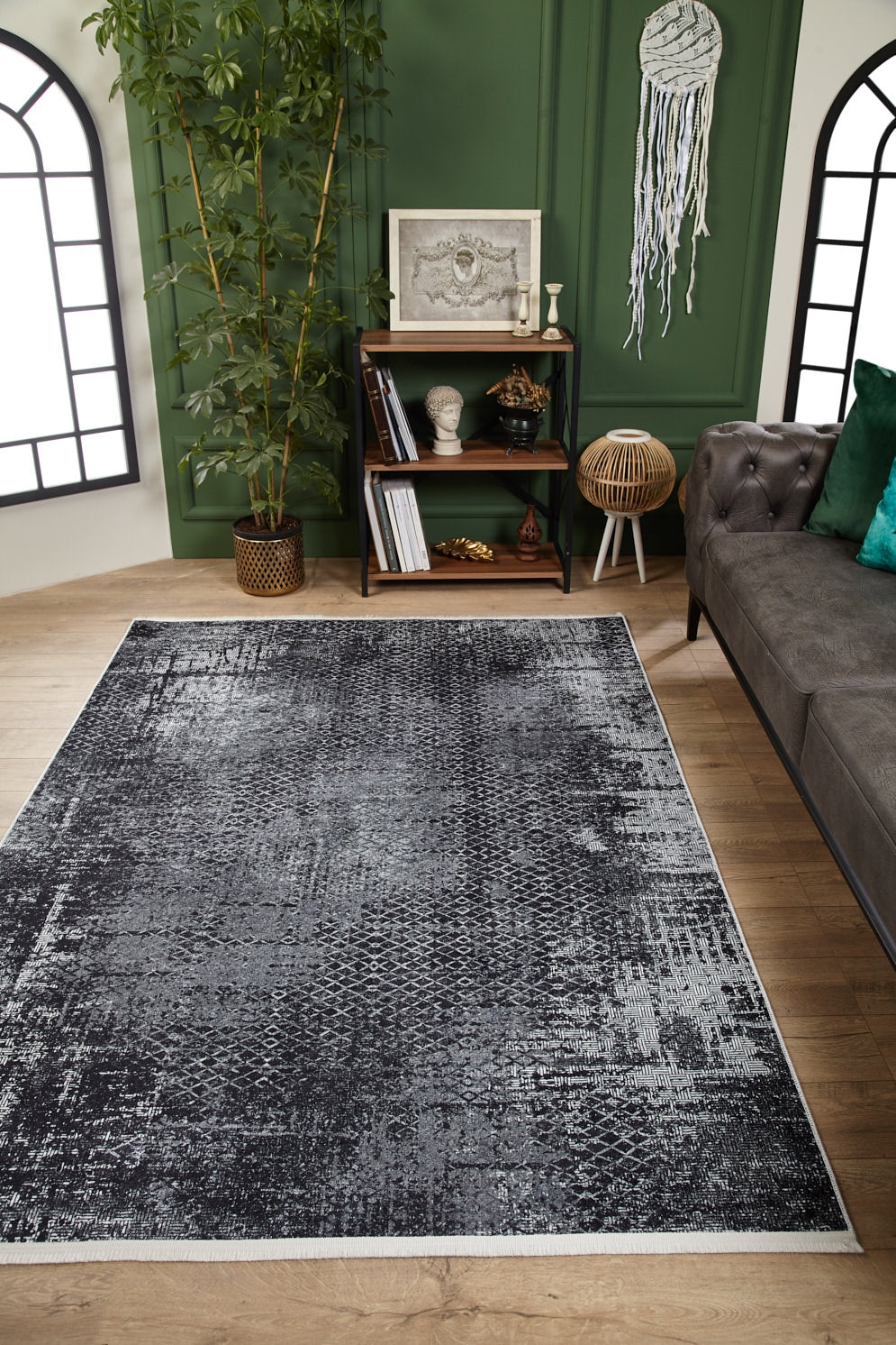 A modern living room featuring the Infinity Garden - Washable Rug - JR1749 (Custom Size) on wooden floors, a dark gray sofa with green cushions, green accent wall, plant, bookshelf, and dreamcatcher—stylish and ideal for high-traffic areas.