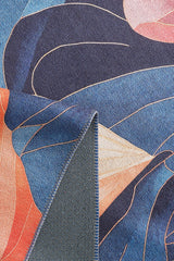 A close-up of the Iris Abstract Floral Washable Rug - MDJ009 shows vibrant blue, orange, pink, and beige abstract shapes. A folded corner reveals its textured blue and gray reverse side on this eye-catching flat-weave rug.