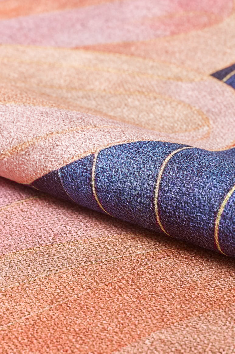 A close-up of the Iris Abstract Floral Washable Rug (MDJ009) reveals its textured flat weave, vibrant pink, peach, and dark blue wavy patterns with thin gold lines. The rug is partially rolled to highlight its multicolor woven design.