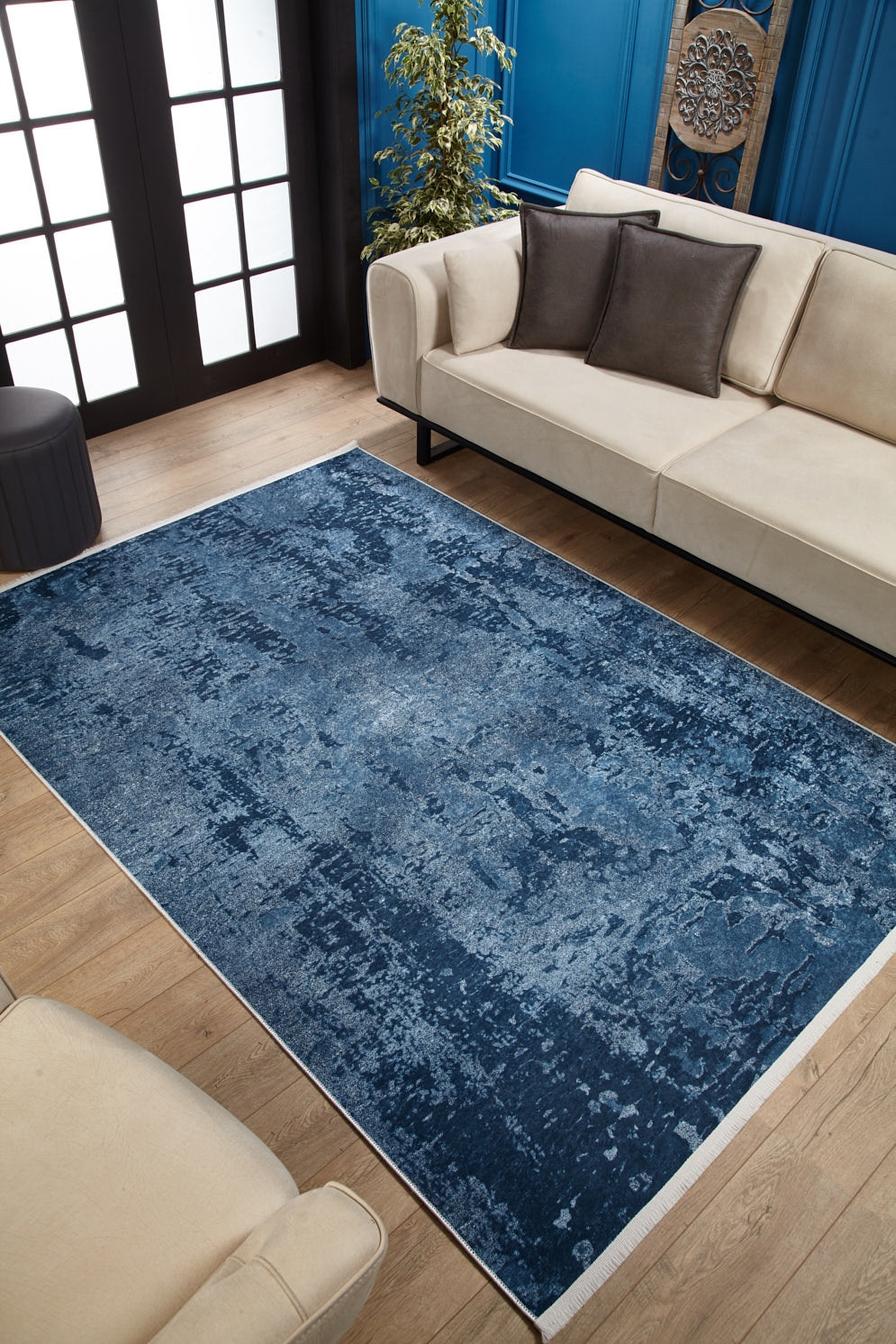 A modern living room features the Ivory Whisper - Washable Rug - JR1628 (Custom Size), ideal for high-traffic areas, on wooden floors with beige sofas, dark pillows, a potted plant, blue paneled walls with art, and French doors in the background.