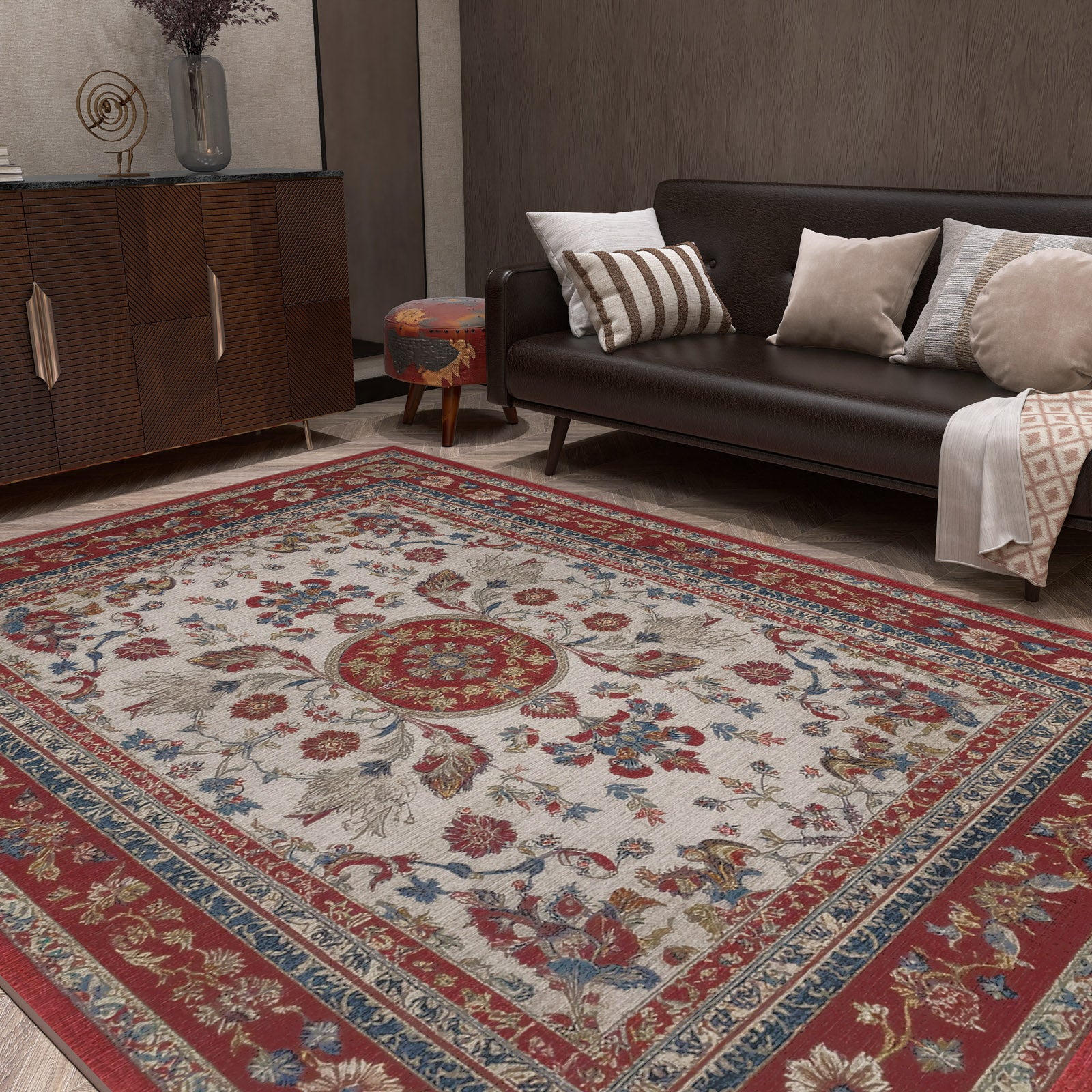 A living room with a dark brown sofa topped with assorted cushions, a wood cabinet, and the Imperial Rose - Washable Rug - JRS023 featuring red, blue, and beige floral patterns. A patterned round ottoman sits near the sofa.