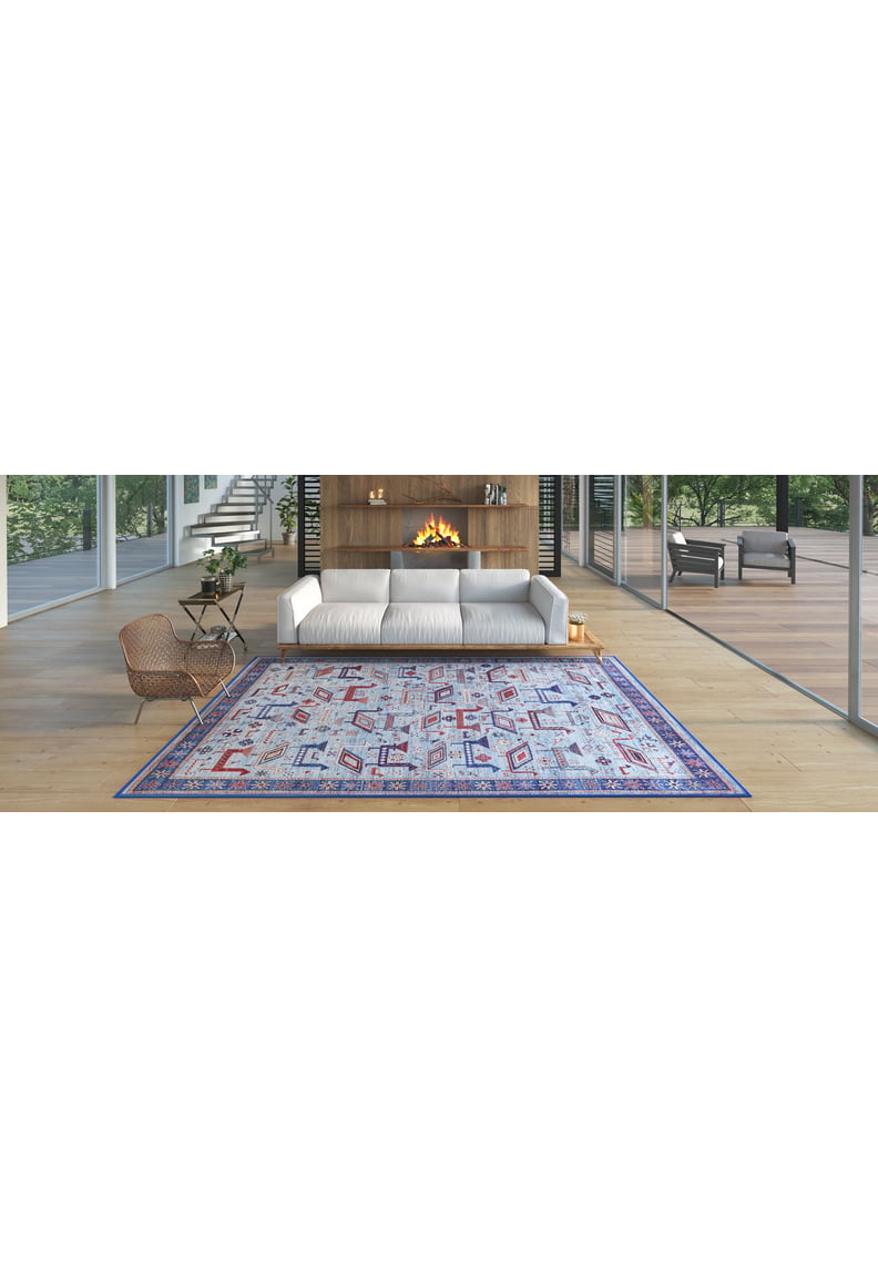 Modern living room with a white sofa, wooden floor, Birdwatcher's Dream Blue Washable Rug - LCC3020 ideal for high-traffic areas, cozy fireplace, and large glass windows overlooking greenery. Wicker chair and small table sit to the left.
