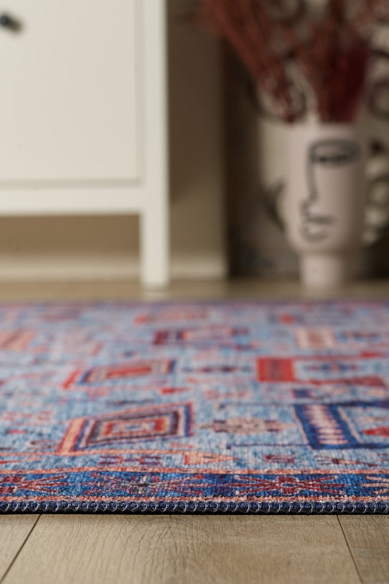 A close-up of the Birdwatcher's Dream Blue Washable Rug - LCC3020 on a wooden floor, with a white cabinet and an abstract face vase in the blurred background—ideal for high-traffic areas.