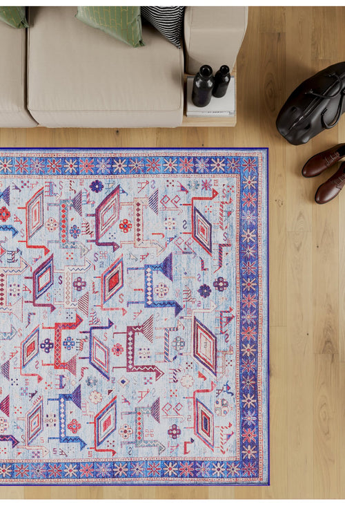 Load image 1 in gallery view. The Birdwatcher's Dream Blue Washable Rug - LCC3020 (Custom Size), with geometric blue, red, and orange patterns and a decorative border, lies on a light wood floor beside a beige sofa, black bag, and brown shoes—ideal for high-traffic areas.