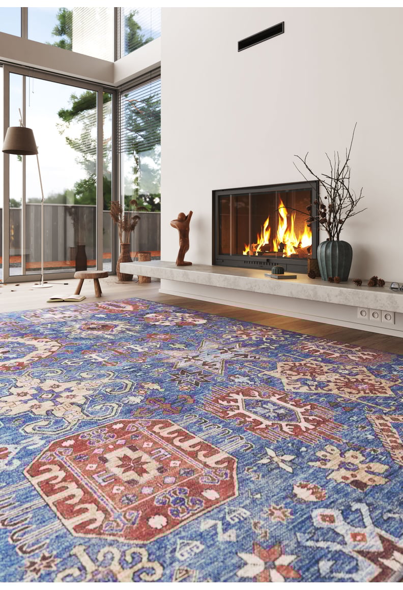 A cozy living room with a lit fireplace, modern decor, floor-to-ceiling windows, and the Kaleidos Authentic Blue Washable Rug—ideal for high-traffic spaces. Sculptures and a vase with branches sit on the mantel.