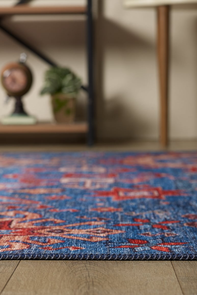 A close-up of the Kaleidos Authentic Blue Washable Rug - LCC3028 (Custom Size) with red patterns on a wooden floor, ideal for high-traffic spaces, featuring blurred shelves, decor, a plant, and a globe in the background.