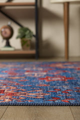 A close-up of the Kaleidos Authentic Blue Washable Rug - LCC3028 (Custom Size) with red patterns on a wooden floor, ideal for high-traffic spaces, featuring blurred shelves, decor, a plant, and a globe in the background.