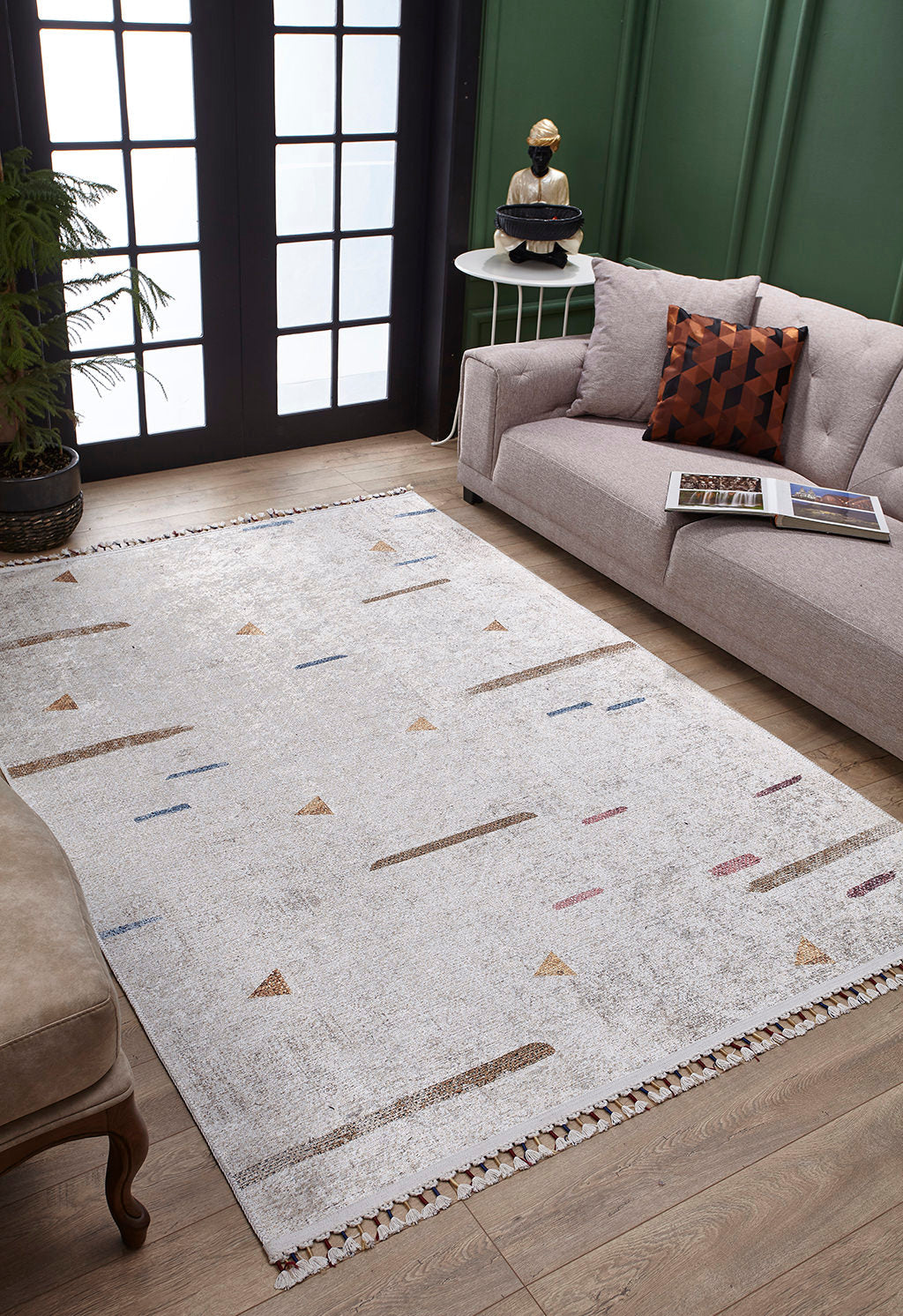 A modern living room features the Kaleidoscope Dreams - Washable Rug - JR5055 in light gray, a beige sofa with a patterned cushion, a small round side table, wooden floors, large glass doors, and green paneled walls for an allergy-sensitive space.
