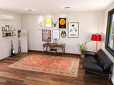A modern living room with wooden floors, the Legacy Wreath - Washable Rug - JRN1024, a black leather sofa, desk with chairs, wall art, shelves of books, a guitar, red floor lamp, mirror, plant, and a large window.