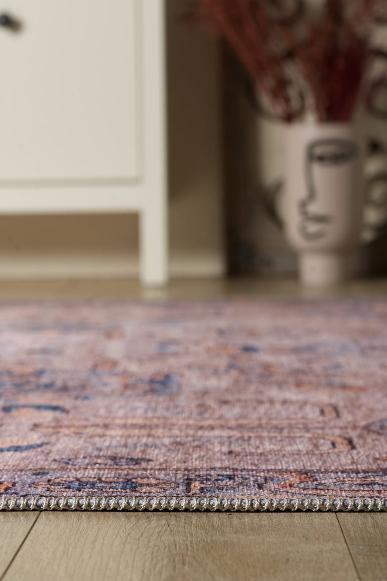 A close-up of the Luminary Beige Gray Washable Rug (LCC3023) on a wooden floor, with part of a white cabinet and a decorative vase with an abstract face design visible in the blurred background.