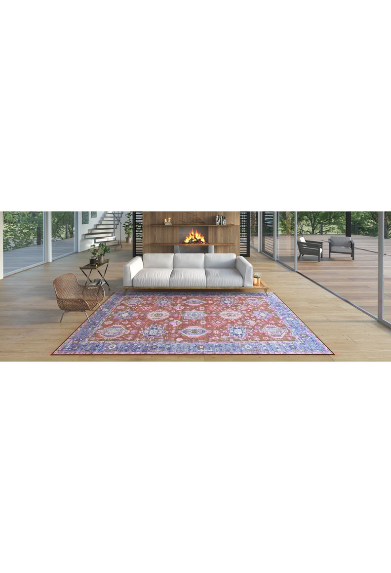 A modern living room features a white sofa, a lit fireplace in a wooden wall, a woven chair, and the Lunavale Red Blue Washable Rug - LCC3007 with colorful patterns on light wood floors, with large windows showcasing trees outside.