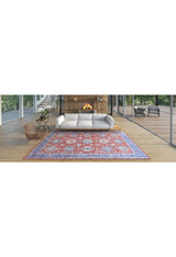 A modern living room features a white sofa, a lit fireplace in a wooden wall, a woven chair, and the Lunavale Red Blue Washable Rug - LCC3007 with colorful patterns on light wood floors, with large windows showcasing trees outside.