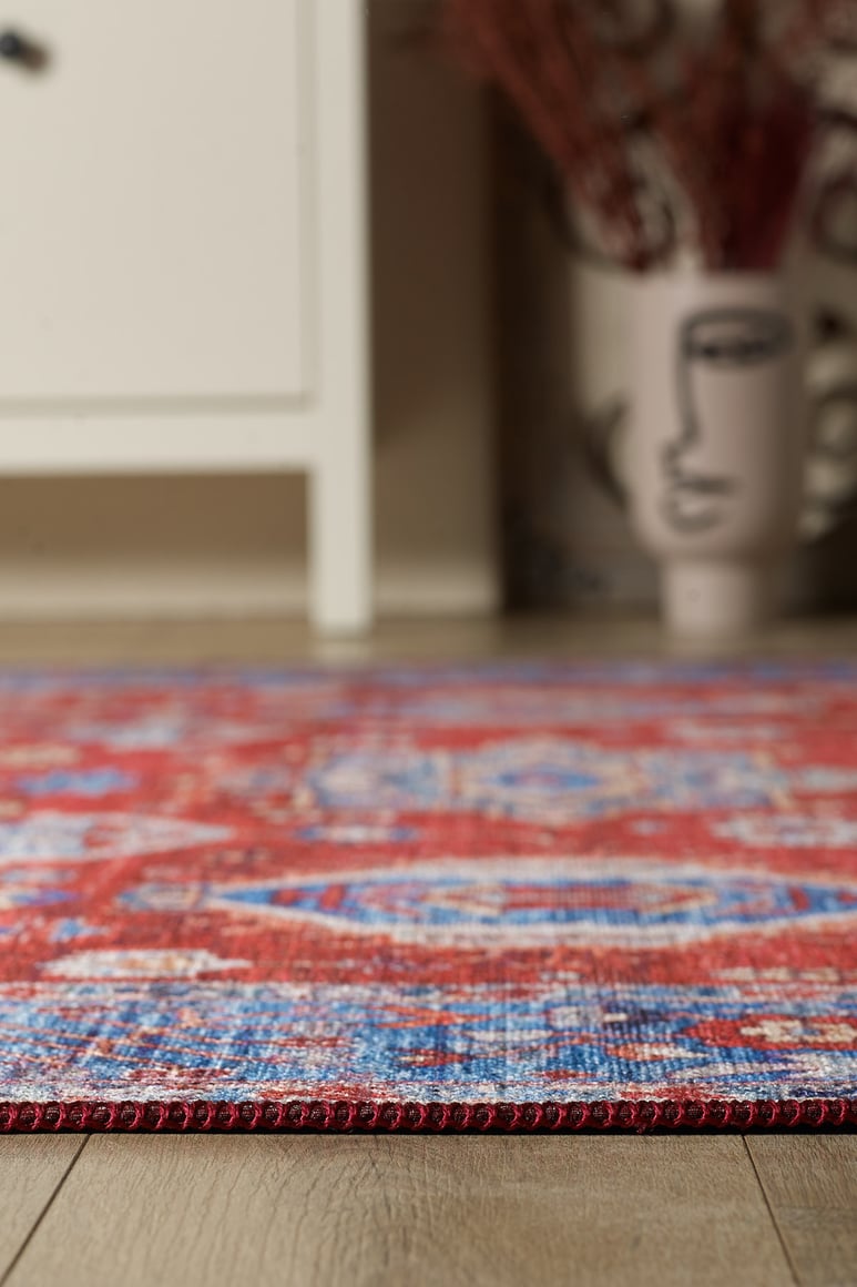 A close-up of the Lunavale Red Blue Washable Rug - LCC3007, designed for high-traffic areas, displays its blue patterns on a wooden floor with a white cabinet and face-design vase blurred in the background.