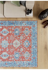 The Lunavale Red Blue Washable Rug - LCC3007 (Custom Size), featuring red, blue, and cream geometric patterns, sits on a light wooden floor beside a beige sofa—ideal for high-traffic areas. Shoes, a bag, and water bottles rest nearby.